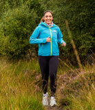 Woman running outdoors in a blue jacket and black leggings with greenery in the background