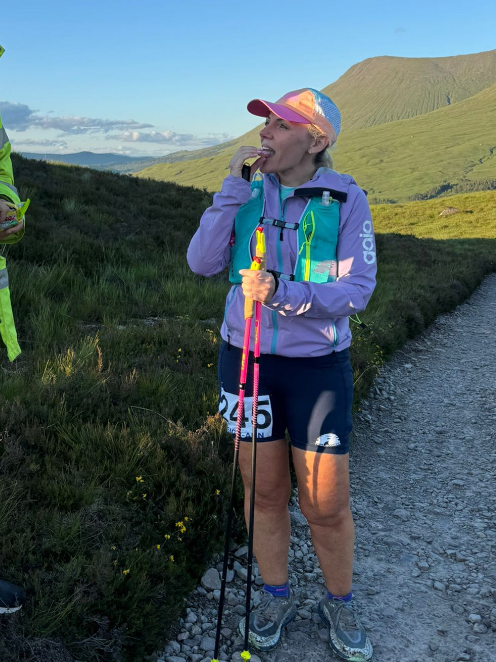 A man in a bright yellow high-visibility jacket stands next to a woman in trail running gear on a dirt path in a mountainous outdoor setting under a clear blue sky.