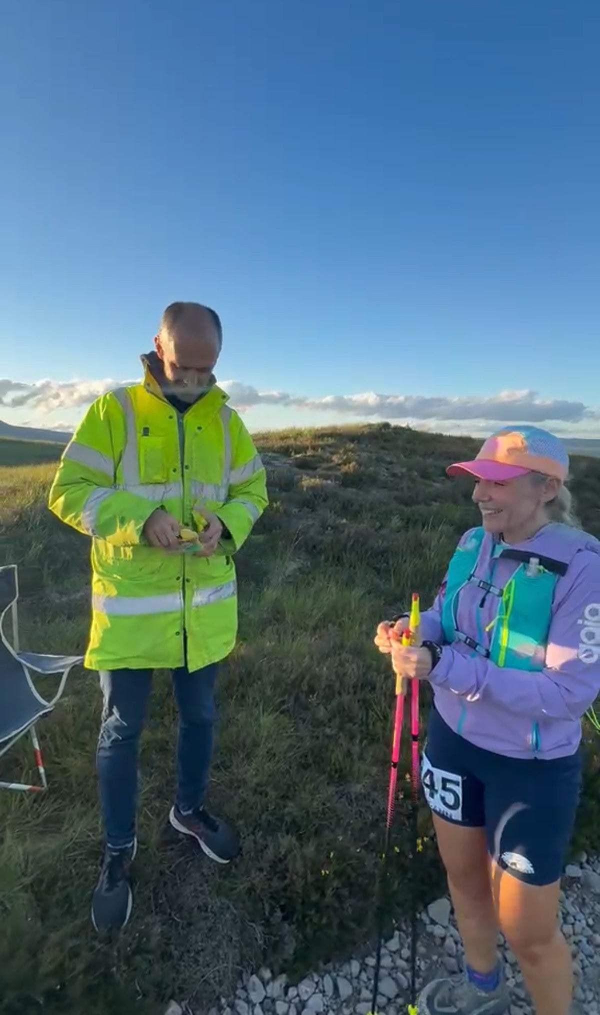 Two people outdoors on a grassy hillside under a blue sky. The person on the left wears a bright high-visibility yellow-green jacket and dark pants, while the person on the right wears a purple top, dark shorts, and a pink cap, holding trekking poles.
