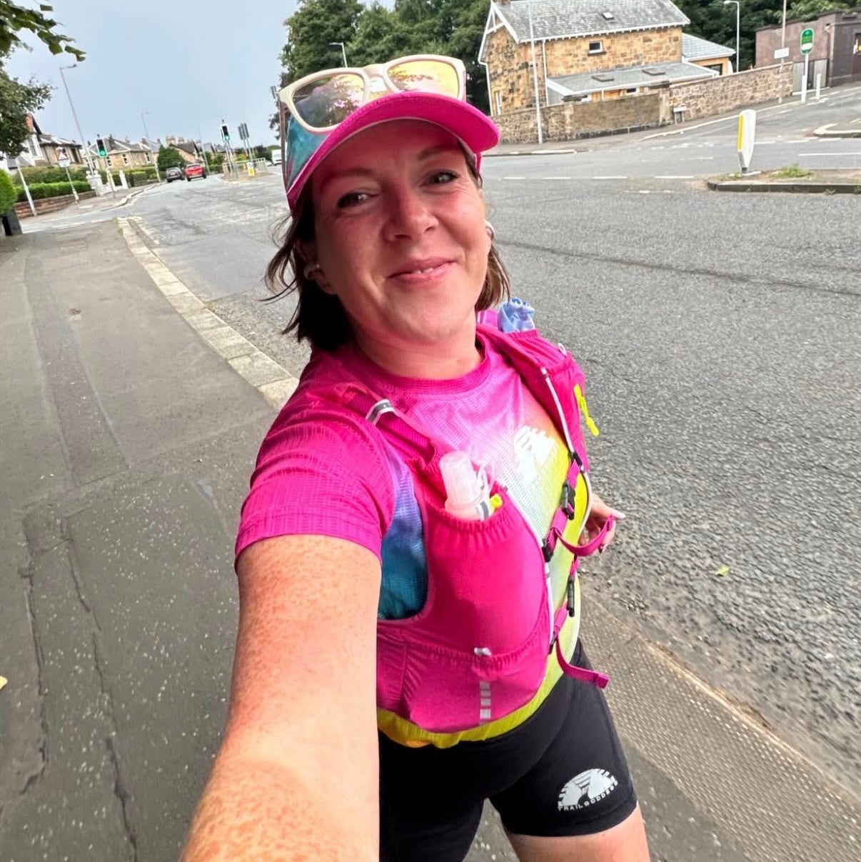 A woman taking a selfie outdoors on a road, wearing a bright pink trail running vest, black running shorts, and a pink cap with sunglasses. The photo has a social media story overlay with text.